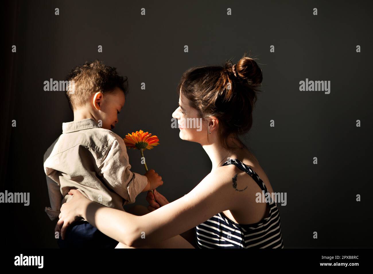 Vero momento elegante bambino Son dà madre gerbera fiore per felice madre giorno contro muro scuro. La mamma ama il bambino. La vita domestica autentica, la luce dura, s Foto Stock