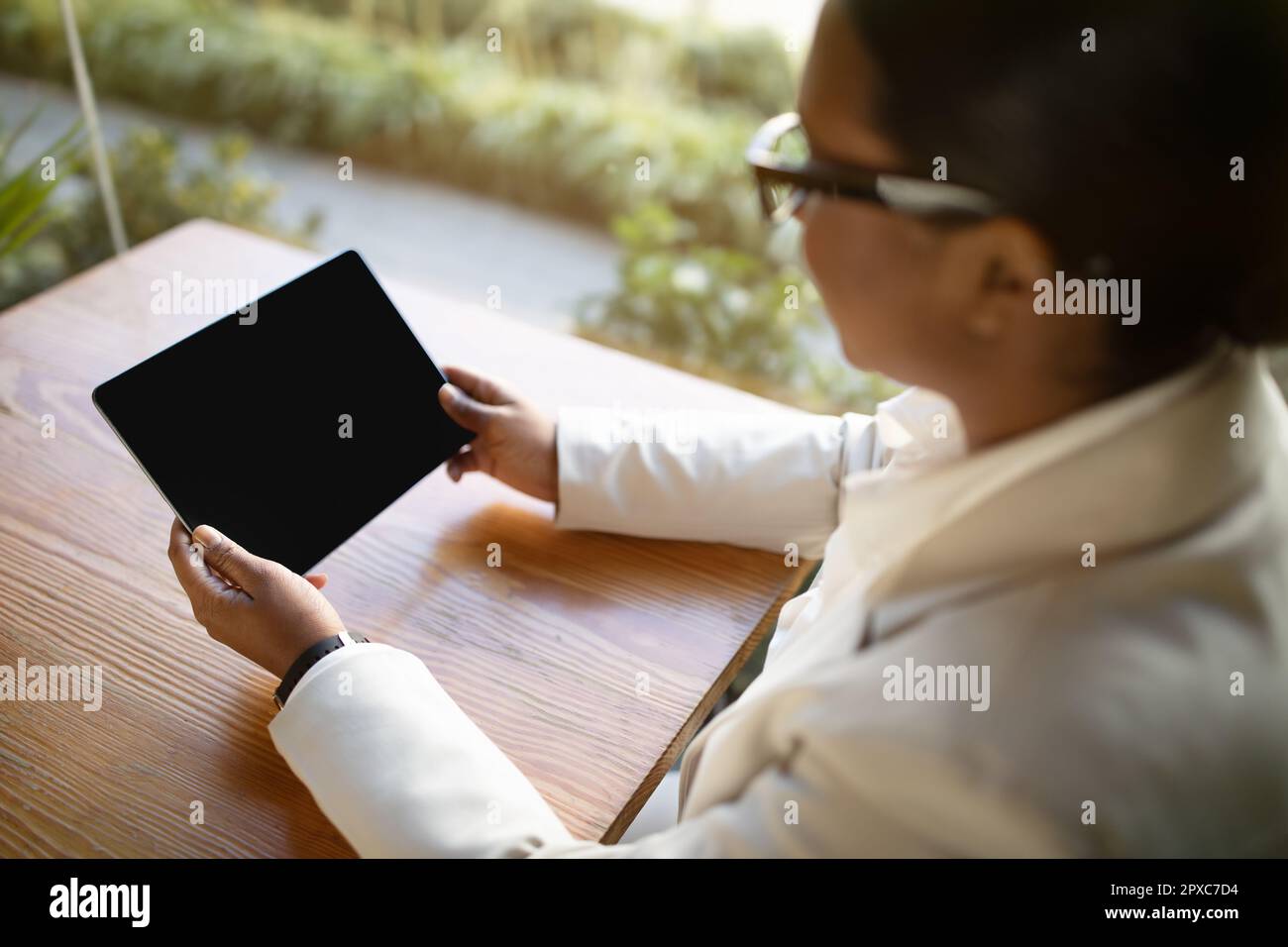 Giovane donna nera in tuta bianca e bicchieri siede al tavolo di legno, guarda il video su un tablet a schermo vuoto Foto Stock