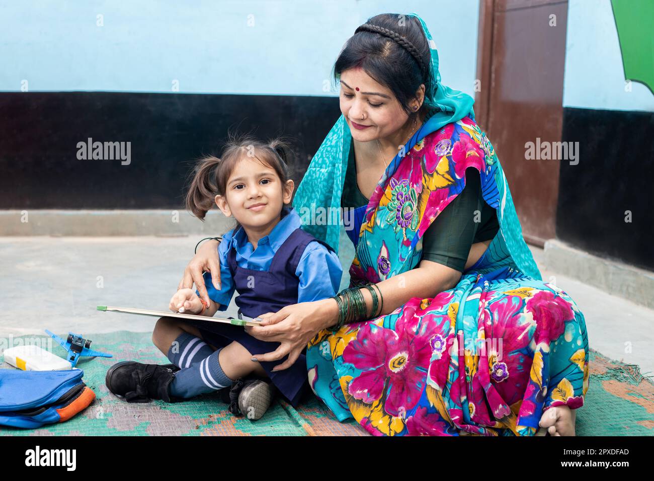 Un giovane insegnante indiano aiuta una bambina studentessa a scrivere su una tavola di ardesia mentre si siede sul pavimento. Bambino che indossa l'uniforme scolastica che studia al governo sc Foto Stock