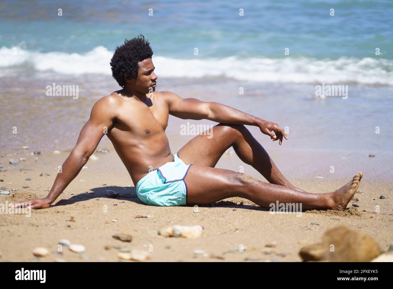 Uomo nero in costume da bagno seduto sulla spiaggia di sabbia Foto Stock