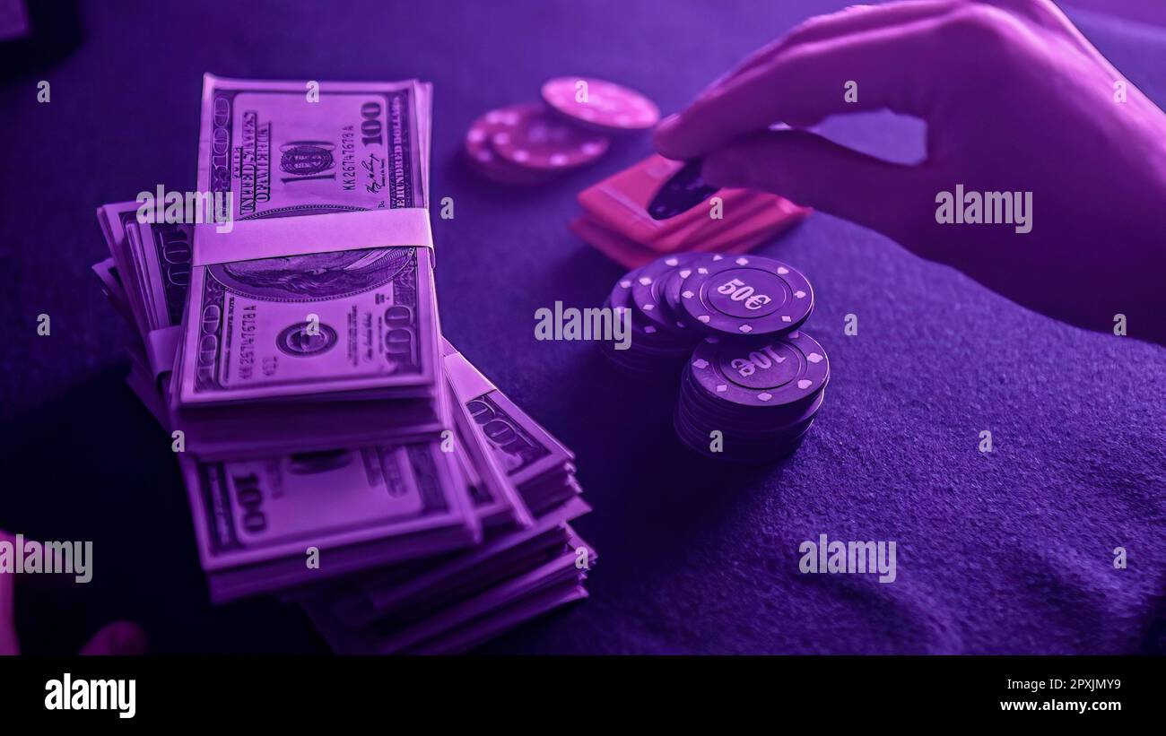 Pila di chip di poker in una gamma di colori e una bacchetta di dollari sul tavolo da gioco in un casinò scarsamente illuminato. Le fiche e i soldi suggeriscono un gioco di poker Foto Stock