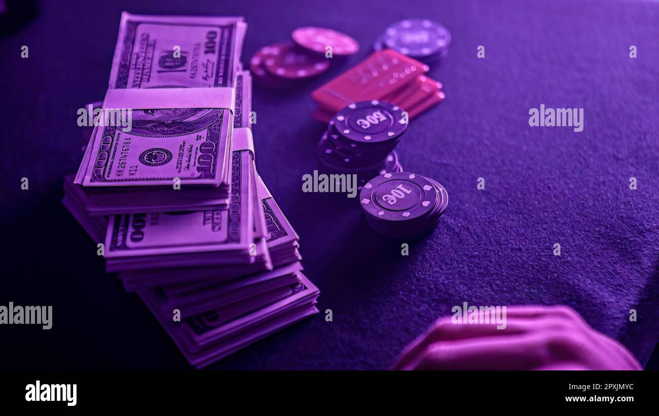 Pila di chip di poker in una gamma di colori e una bacchetta di dollari sul tavolo da gioco in un casinò scarsamente illuminato. Le fiche e i soldi suggeriscono un gioco di poker Foto Stock