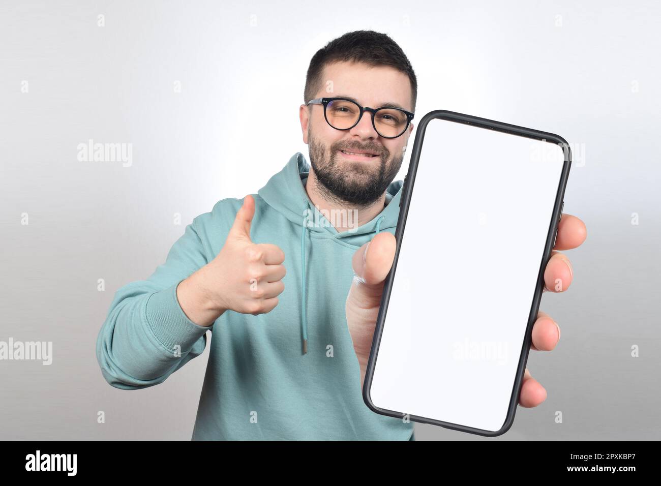 un giovane uomo con gli occhiali e la barba mostra un telefono con uno schermo bianco vuoto Foto Stock