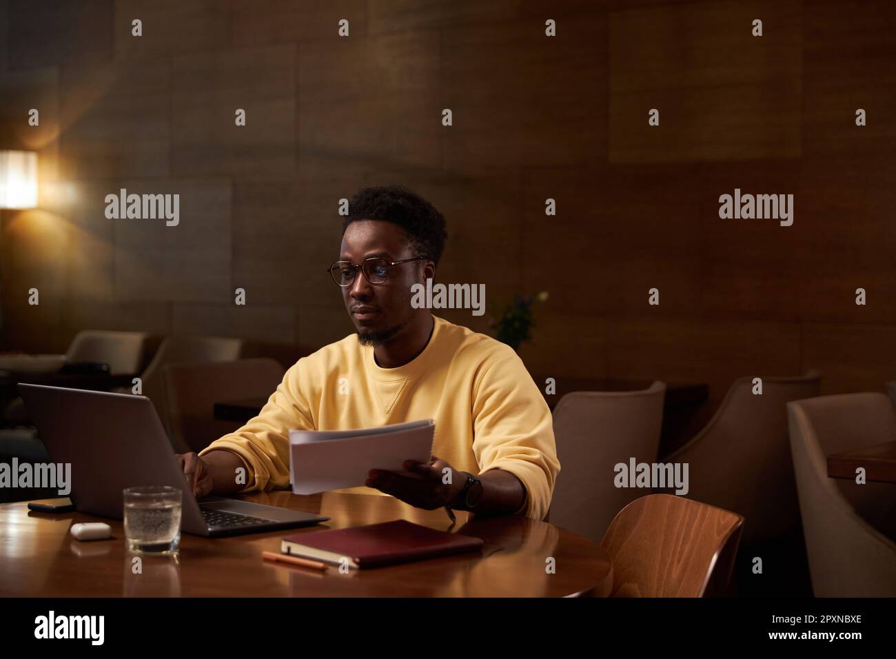 Giovane uomo d'affari afroamericano che lavora su un computer portatile con documenti mentre si siede al tavolo nel ristorante Foto Stock