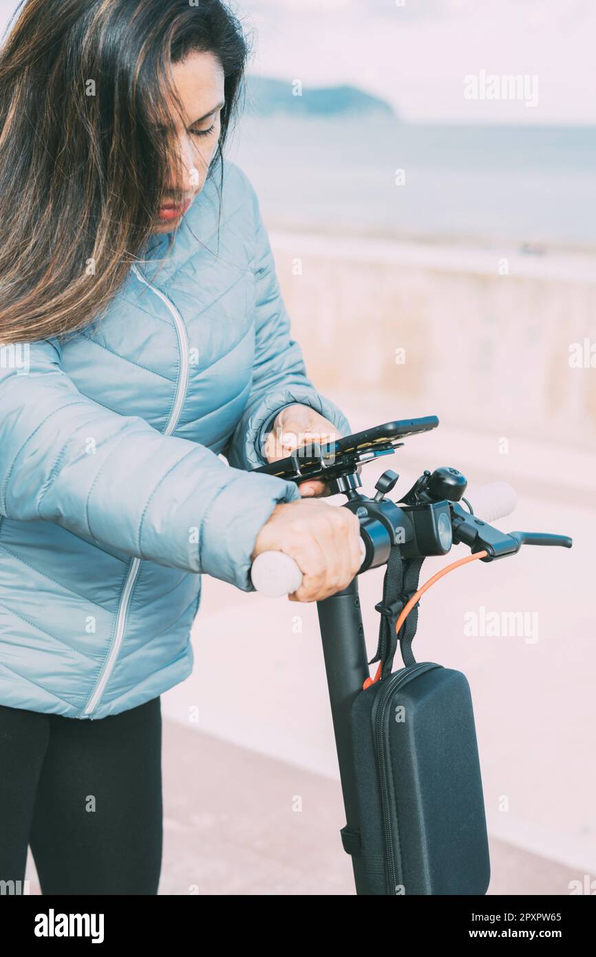 Vista laterale della bella donna latina in sella al suo scooter motorizzato in città, attivazione di app per smartphone, concetto di mobilità trasporto sostenibile, zero Foto Stock