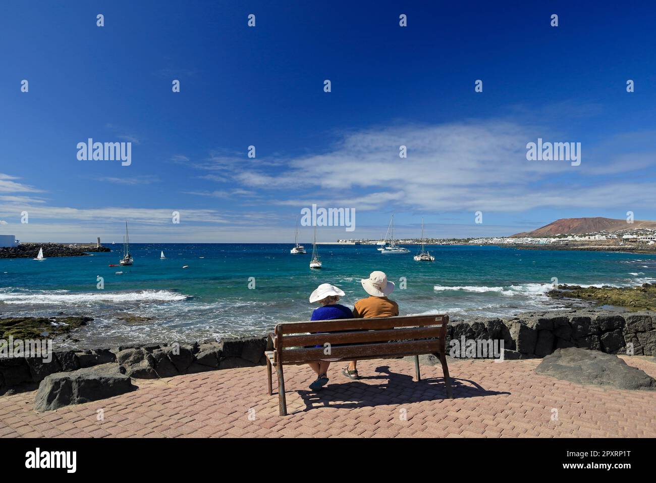Coppia matura seduta su panchina che si affaccia su una bella vista sul mare, barche e onde. Lanzarote ha preso febbraio 2023. Foto Stock