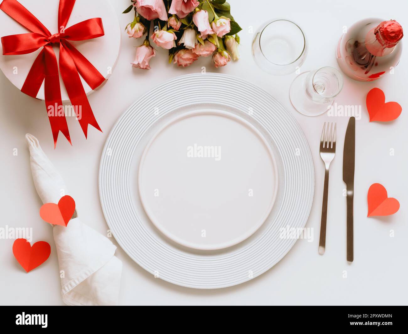 Concetto di tablesettings di San Valentino su sfondo bianco. Piatti bianchi vuoti, posate dorate, bottiglia di vino rosa, bicchieri, bouqet di fiori, confezione regalo. Copia Foto Stock