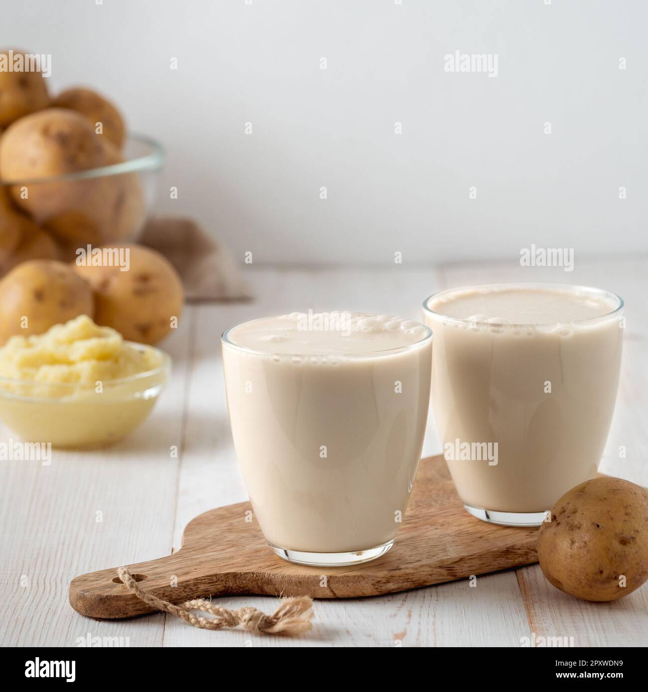 Latte di patate in bicchieri su sfondo bianco di legno. Versare il latte vegano in vetro, con purea di patate e tuberi di patate sullo sfondo. Spazio di copia. Ma. Iniziale Foto Stock