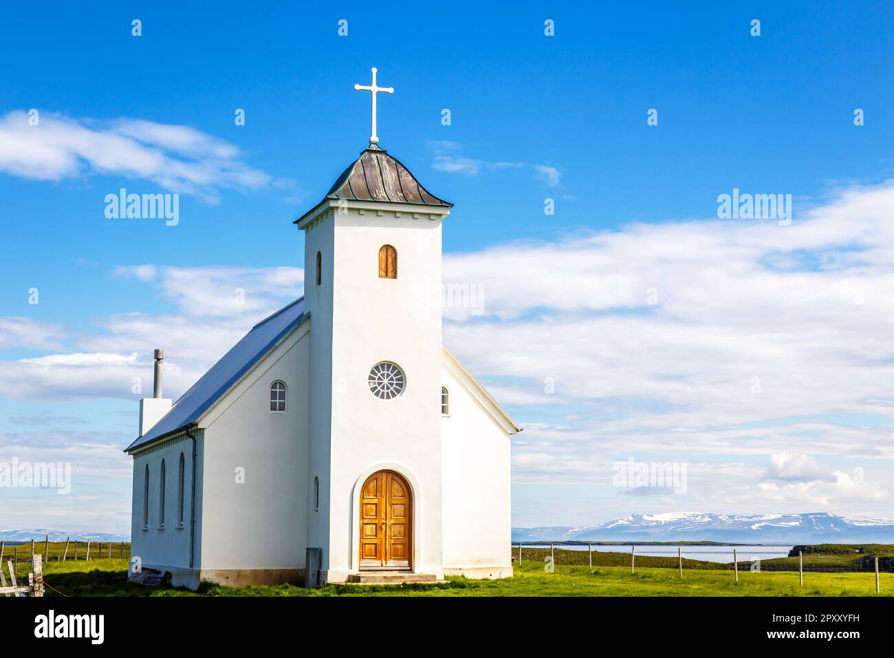 Flateyjarkirkja bianca chiesa luterana con prato in primo piano e fiordo di mare con cielo blu e montagne sullo sfondo, Flatey Island, Islanda Foto Stock