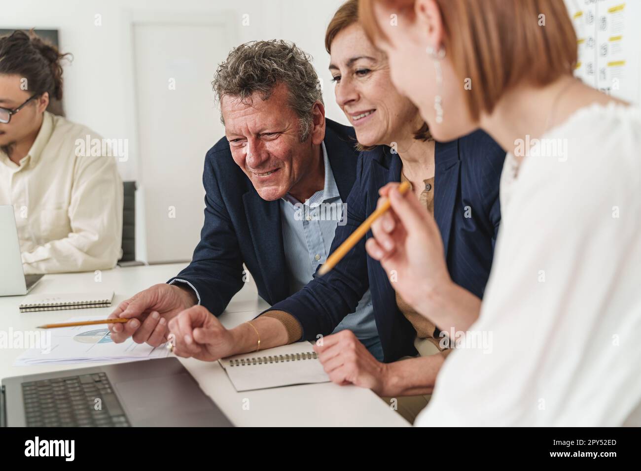 In un ufficio moderno, tre colleghi caucasici vestiti casualmente, due anziani e una giovane donna, si concentrano sullo schermo di un notebook, mentre un giovane asiatico lavora Foto Stock