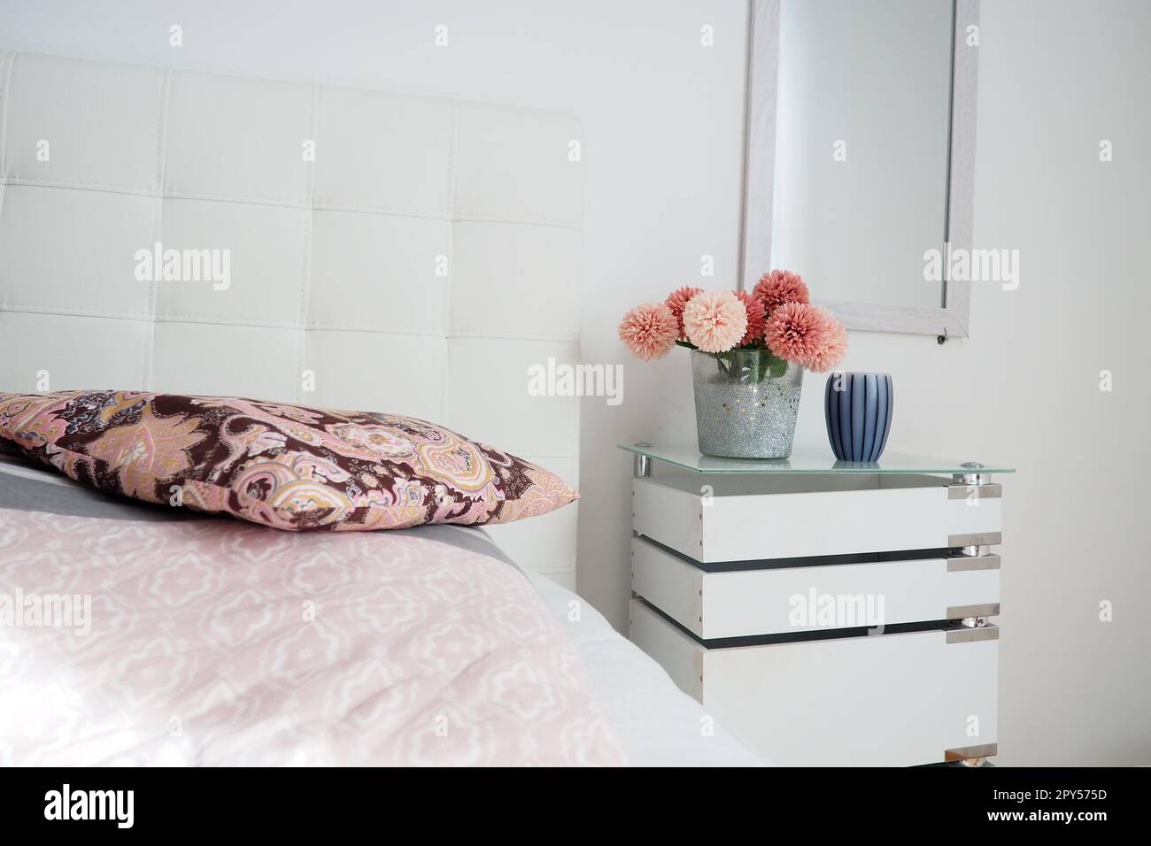 Design interno della camera da letto. Specchio sul muro. Un bouquet di bellissimi fiori rosa su un tavolo bianco in camera da letto. Design degli interni. Letto con testiera imbottita bianca. Cuscino decorativo Foto Stock