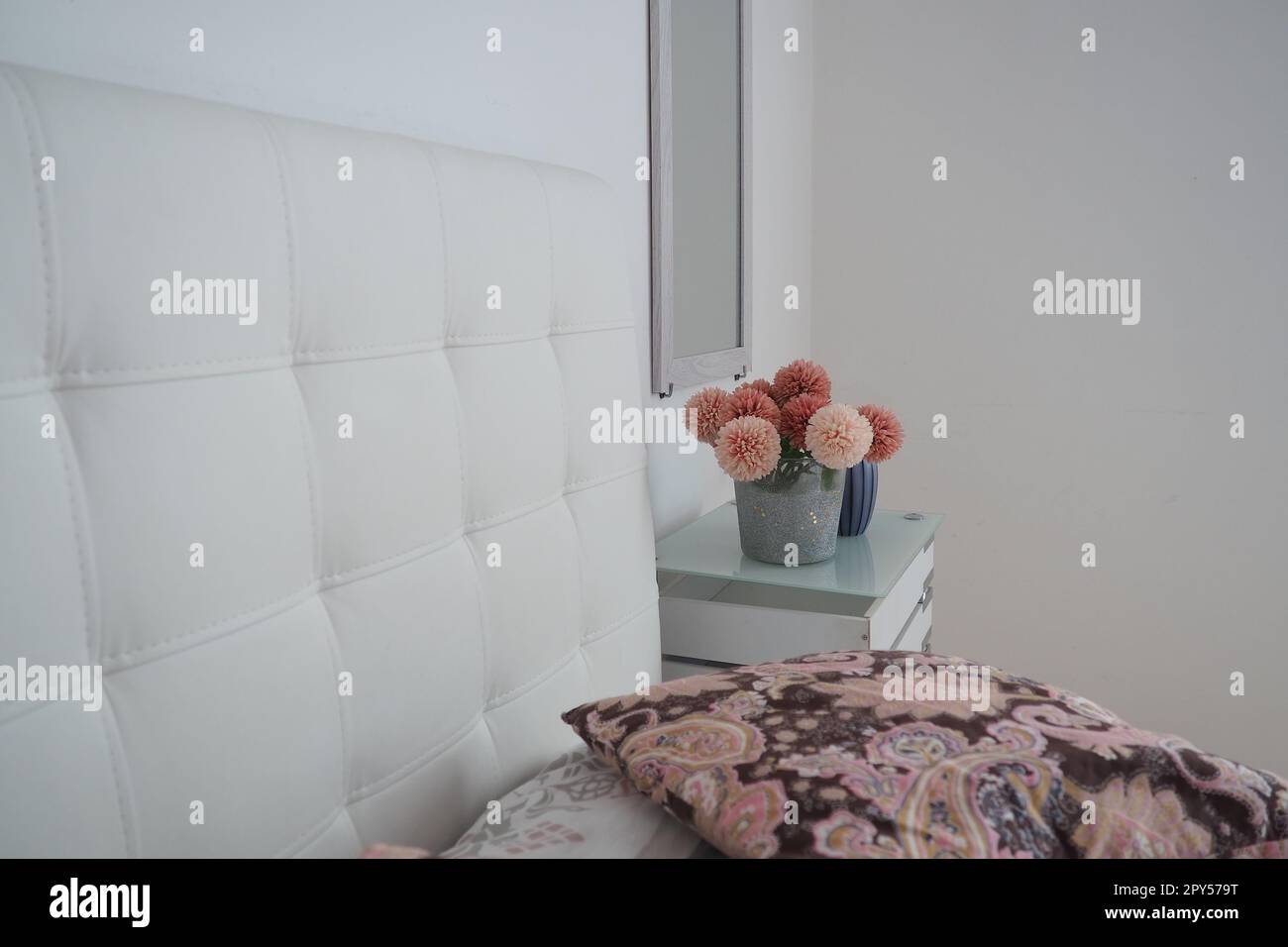 Design interno della camera da letto. Specchio sul muro. Un bouquet di bellissimi fiori rosa su un tavolo bianco in camera da letto. Design degli interni. Letto con testiera imbottita bianca. Cuscino decorativo Foto Stock