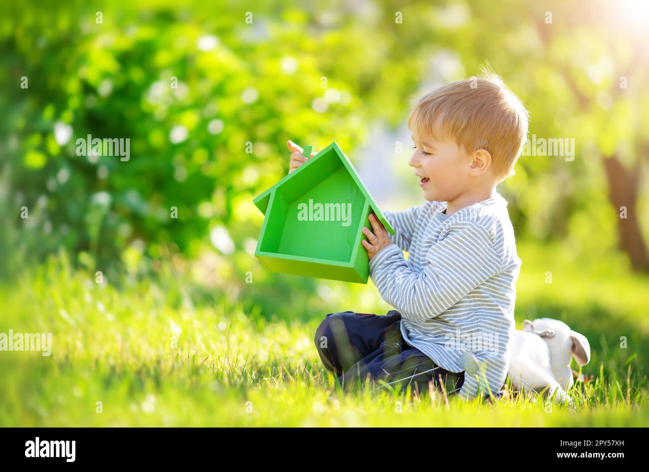 Bambino sorridente seduto sul prato di primavera con il modello della casa verde nelle sue mani. Foto Stock