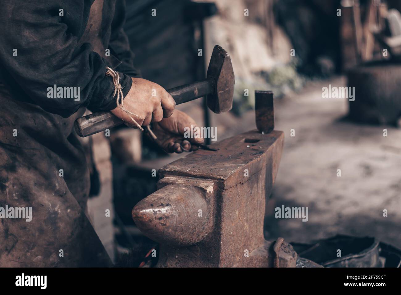 Metallo di lavorazione fabbro con martello su incudine in fucina. Farrier con i vestiti sporchi colpisce il ferro in officina. Lavorazione dei metalli. Foto Stock
