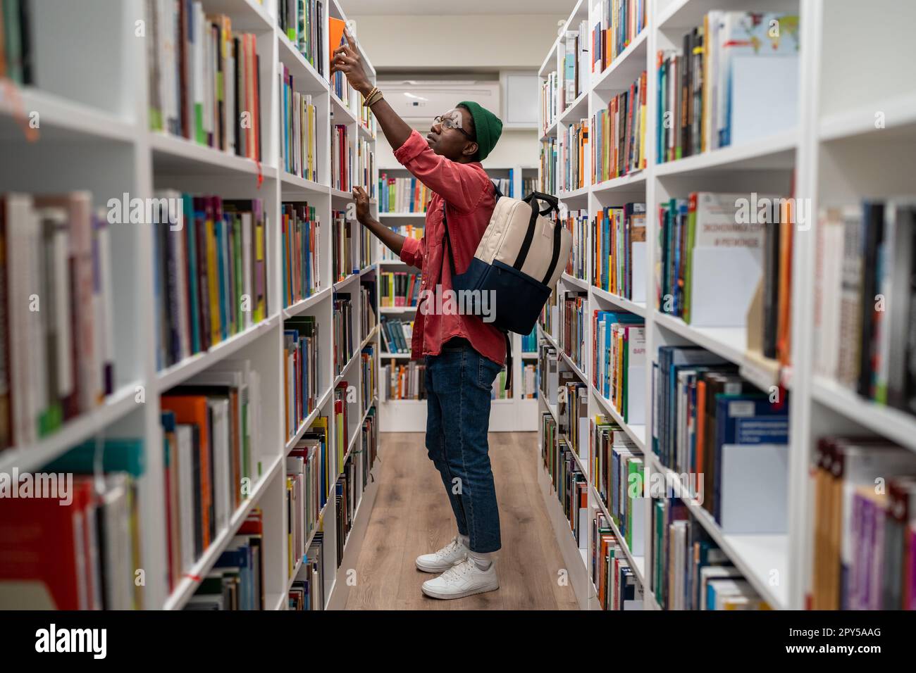 Allievo nerd uomo afroamericano che sceglie il libro nella biblioteca universitaria prendendolo dallo scaffale. Foto Stock
