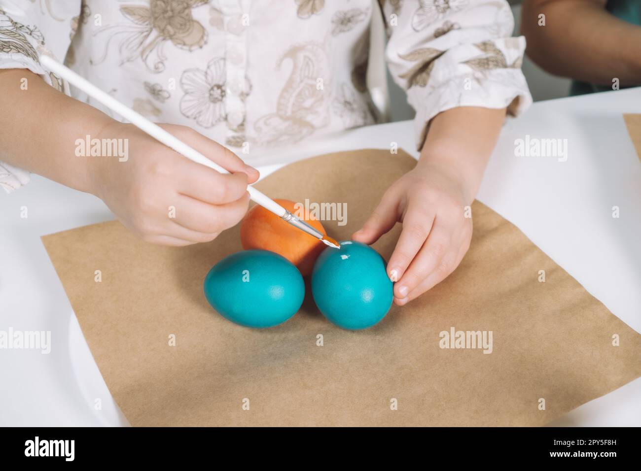 Ritratto ritagliato della mano di una bambina, che dipinge uova di galline pasquali multicolore, tavolo con lastre di carta marrone, pennello Foto Stock