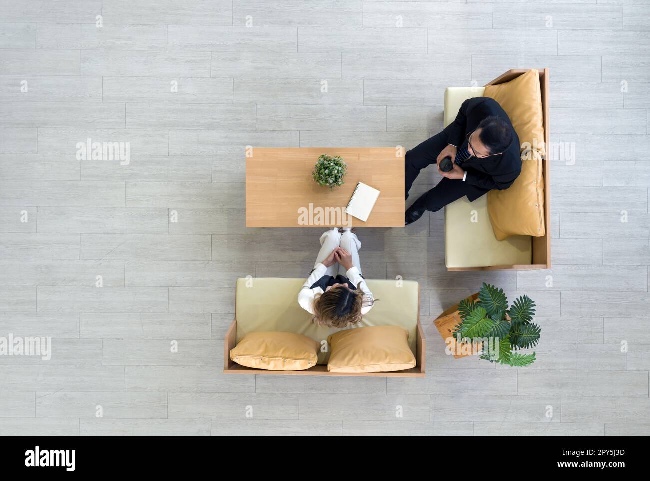 Vista ad alto angolo del moderno spazio di coworking. Giovane donna d'affari asiatica in abito bianco che parla con il senior manager con una tazza di caffè in mano. Vasi di fiori e note-book sono su un tavolo di legno. Foto Stock