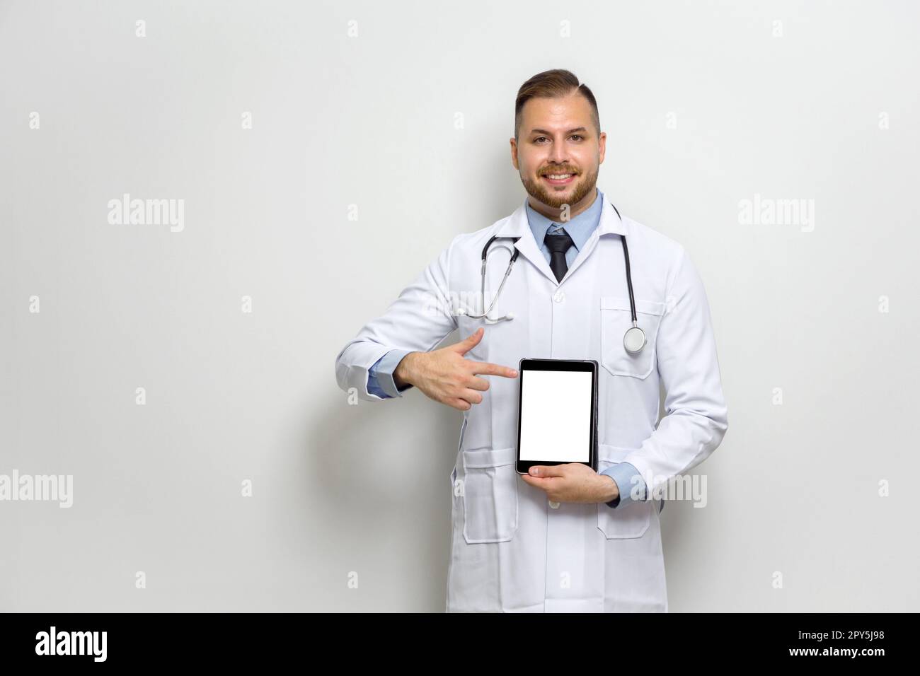 Giovane medico caucasico con capelli corti, baffi e barba vestita di abito bianco e stetoscopio che punta il dito verso il monitor del computer tablet. Posizionatevi di fronte alla parete bianca. Foto Stock