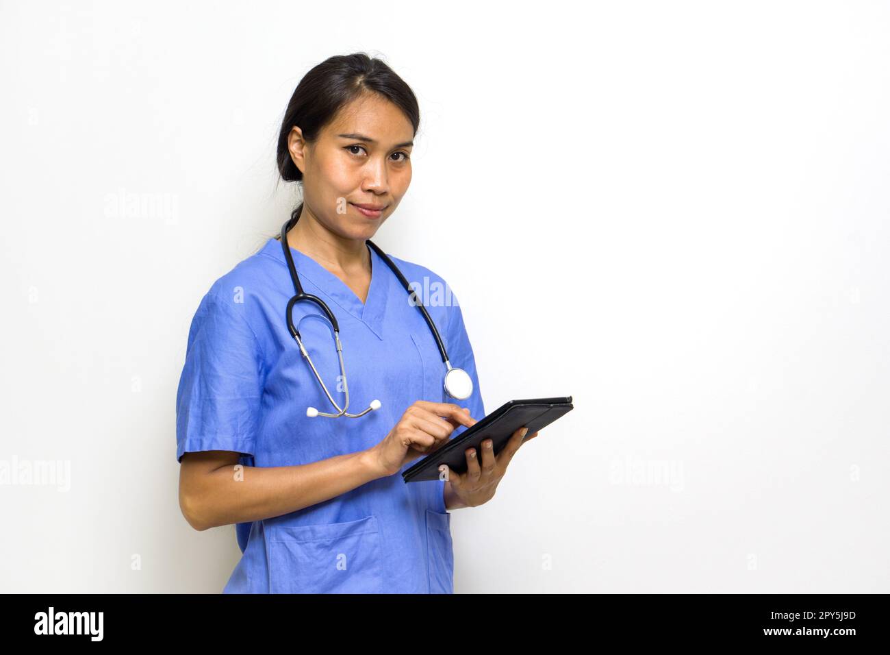 Fisioterapista donna in uniforme blu e stetoscopio a scrivere sul computer tablet. Posizionarsi di fronte alla parete bianca. Foto Stock