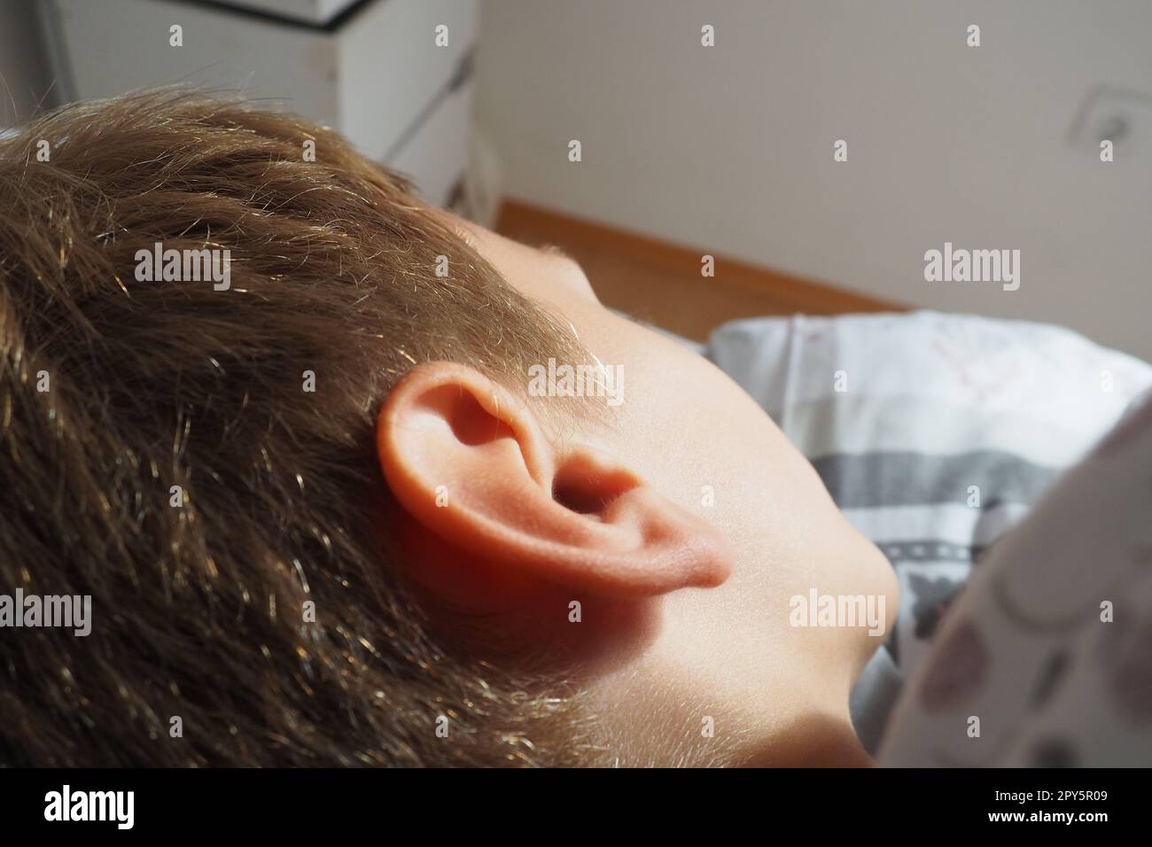 Un bel ragazzo caucasico di 8 anni con i capelli biondi dorme su un letto con una soffice coperta e un cuscino. La luce soffusa del sole mattutino. Primo piano dell'orecchio. Il bambino sta riposando. Sonno sano. Foto Stock