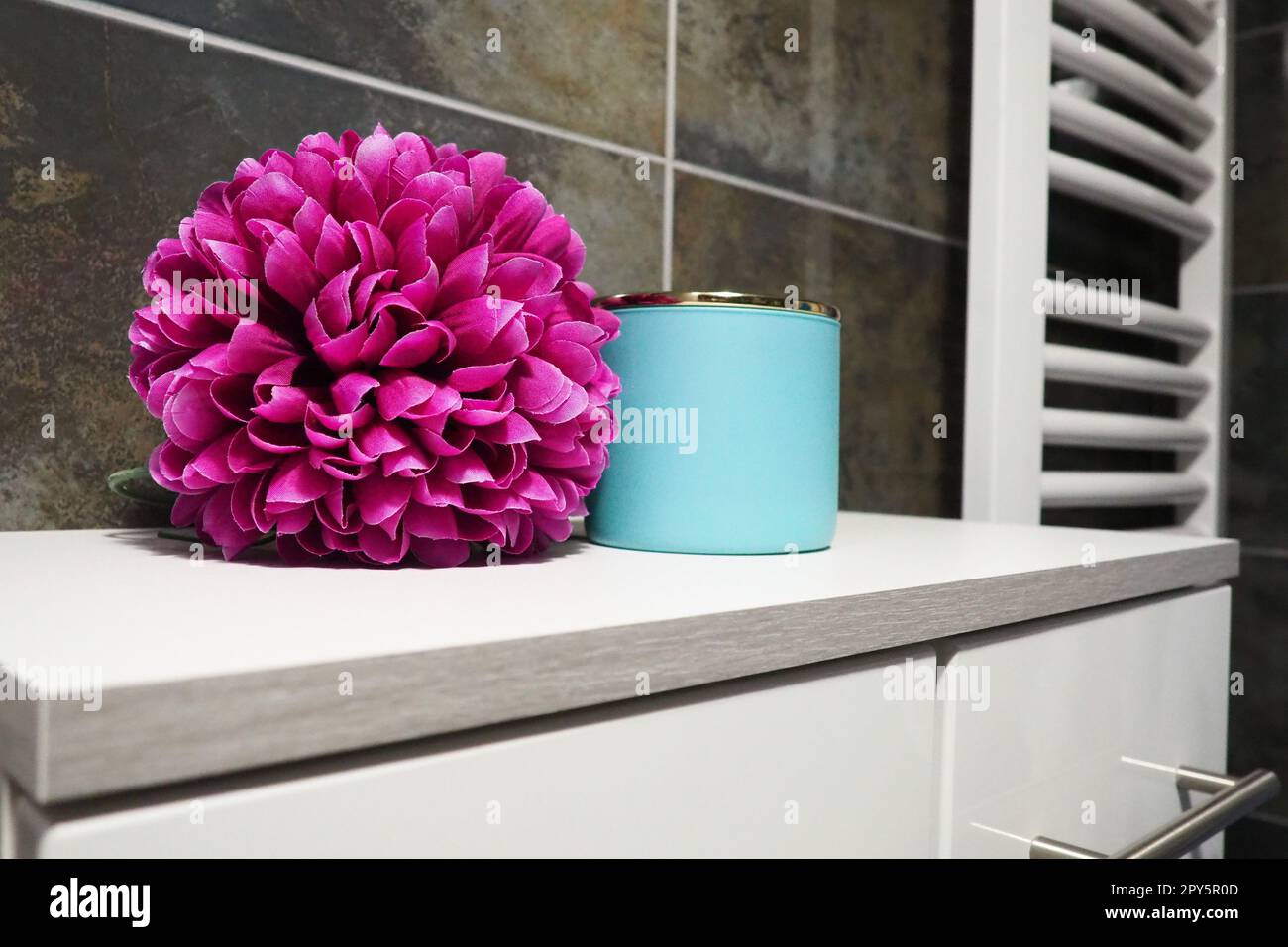 Elementi decorativi del bagno. Un barattolo blu di crema e un doppio fiore artificiale rosa brillante su un ripiano del mobile. Piastrelle nere. Bagno e WC interno. Scaldasalviette per radiatore sulla parete. Foto Stock