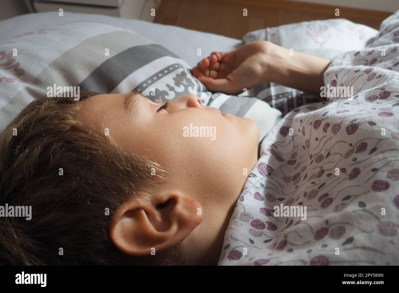 Un bel ragazzo caucasico di 8 anni con i capelli biondi dorme su un letto con una soffice coperta e un cuscino. La luce soffusa del sole mattutino. Primo piano dell'orecchio. Il bambino sta riposando. Sonno sano. Foto Stock