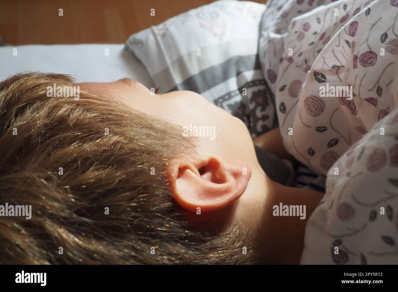 Un bel ragazzo caucasico di 8 anni con i capelli biondi dorme su un letto con una soffice coperta e un cuscino. La luce soffusa del sole mattutino. Primo piano dell'orecchio. Il bambino sta riposando. Sonno sano. Foto Stock