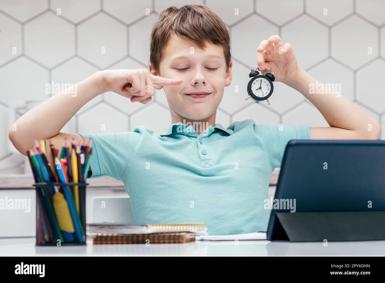 Ritratto di un giovane ragazzo felice con gli occhi chiusi seduto alla scrivania con un tablet digitale, puntando il dito indice a un piccolo orologio. Foto Stock