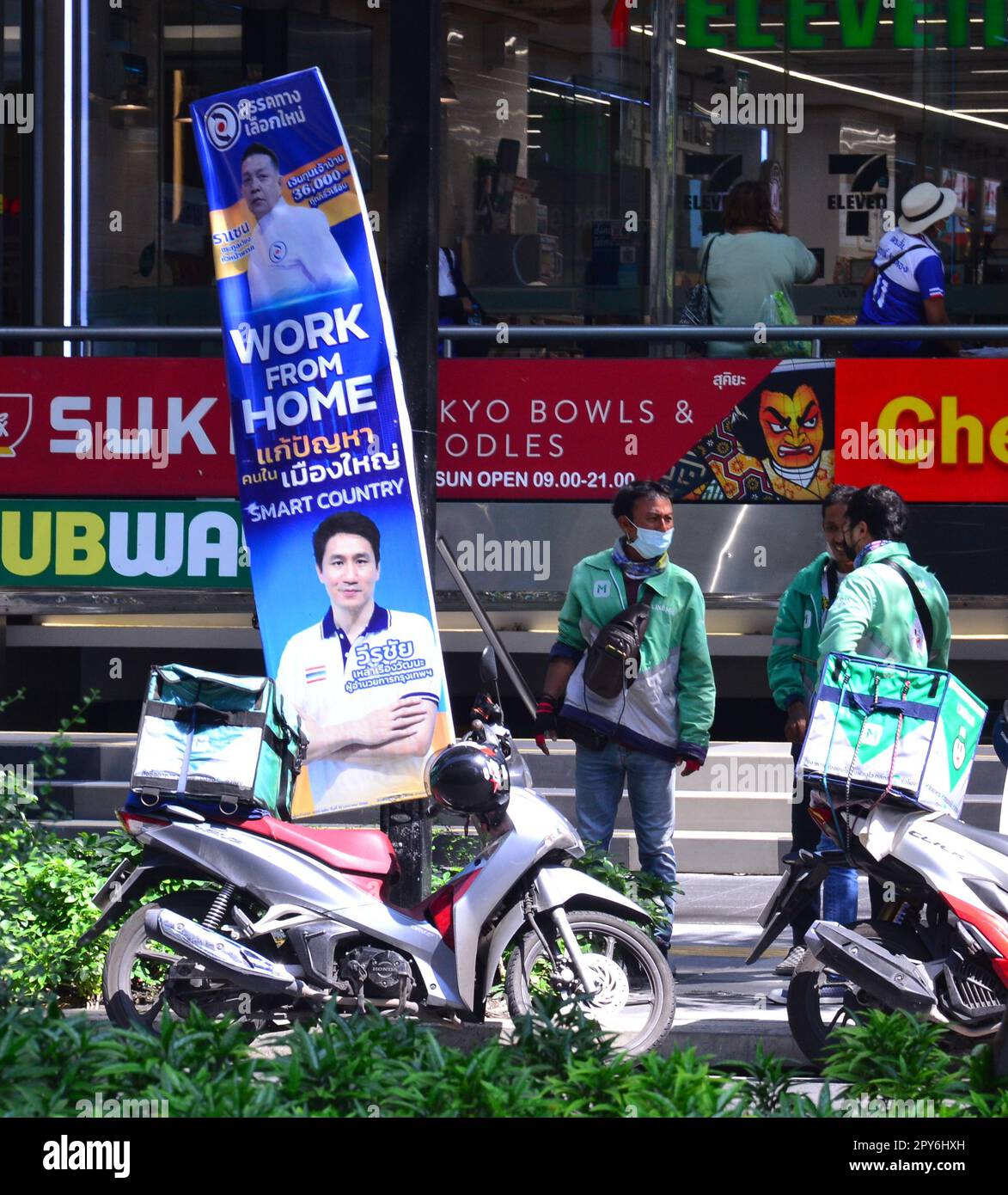 Motociclisti addetti alla consegna dei prodotti alimentari accanto a un cartello elettorale il cui testo include "lavoro da casa". Un candidato alle elezioni utilizza un cartello allegato ai mobili per la strada per pubblicizzare se stesso presso potenziali elettori nel distretto di Silom, nel centro di Bangkok, in Thailandia, nel sud-est asiatico. Le elezioni generali si terranno in Thailandia il 14th maggio 2023, dopo lo scioglimento della 25th Camera dei rappresentanti il 20th marzo 2023. Foto Stock