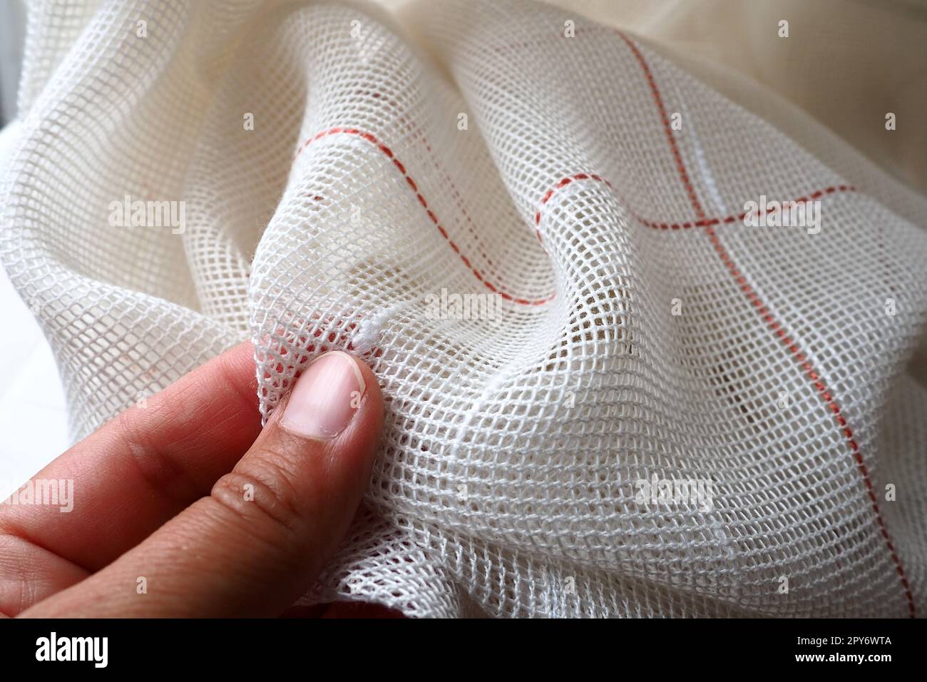 Tende bianche in cotone grossolano o lino fatte di trame di fili sottili. La mano di una donna sente il tessuto. Camera decorativa con finestre. Striscia rossa. Il tessuto è piegato a onde morbide. Interno dell'appartamento. Foto Stock