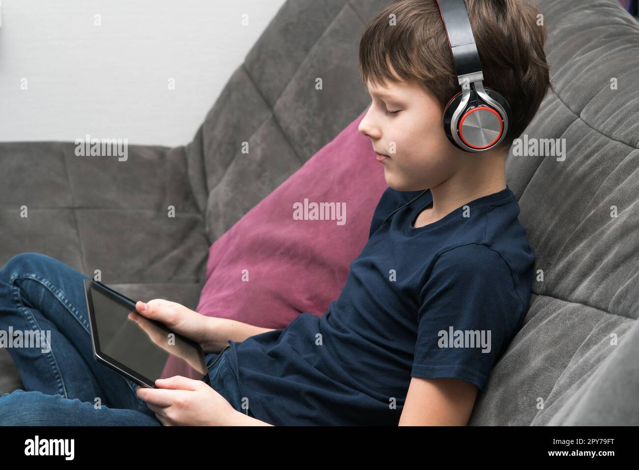 Bambino allegro e spirituale con cuffie che guarda film e ascolta musica con il tablet digitale e il servizio di streaming Foto Stock