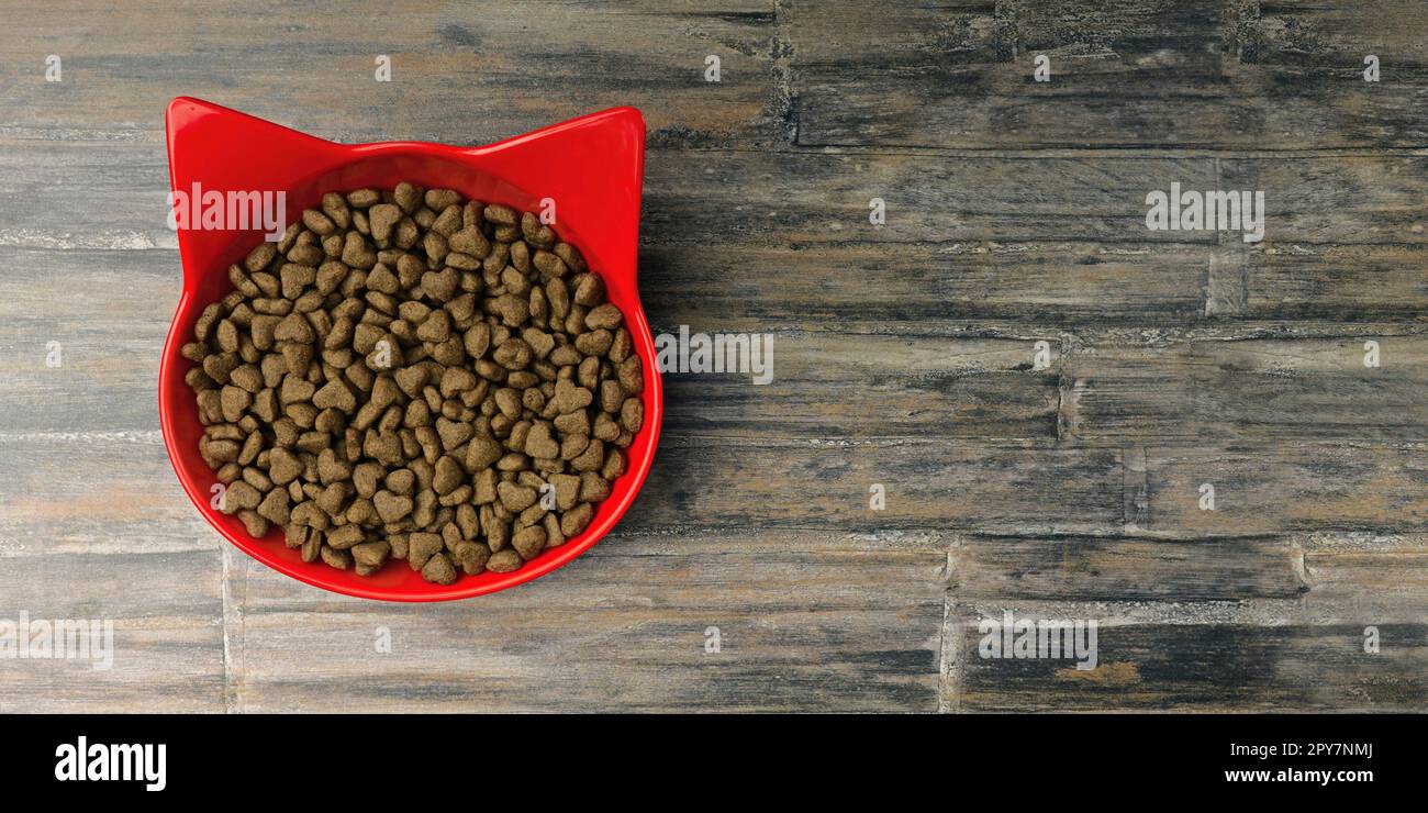 Ciotola rossa a forma di gatto riempita con cibo secco per animali domestici su sfondo rustico. Immagine panoramica con spazio di copia. Foto Stock