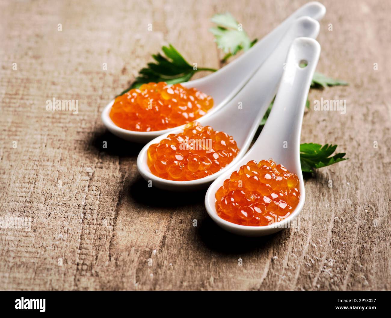 Primo piano del lussuoso caviale rosso in cucchiaio bianco con prezzemolo su fondo di legno. Foto Stock