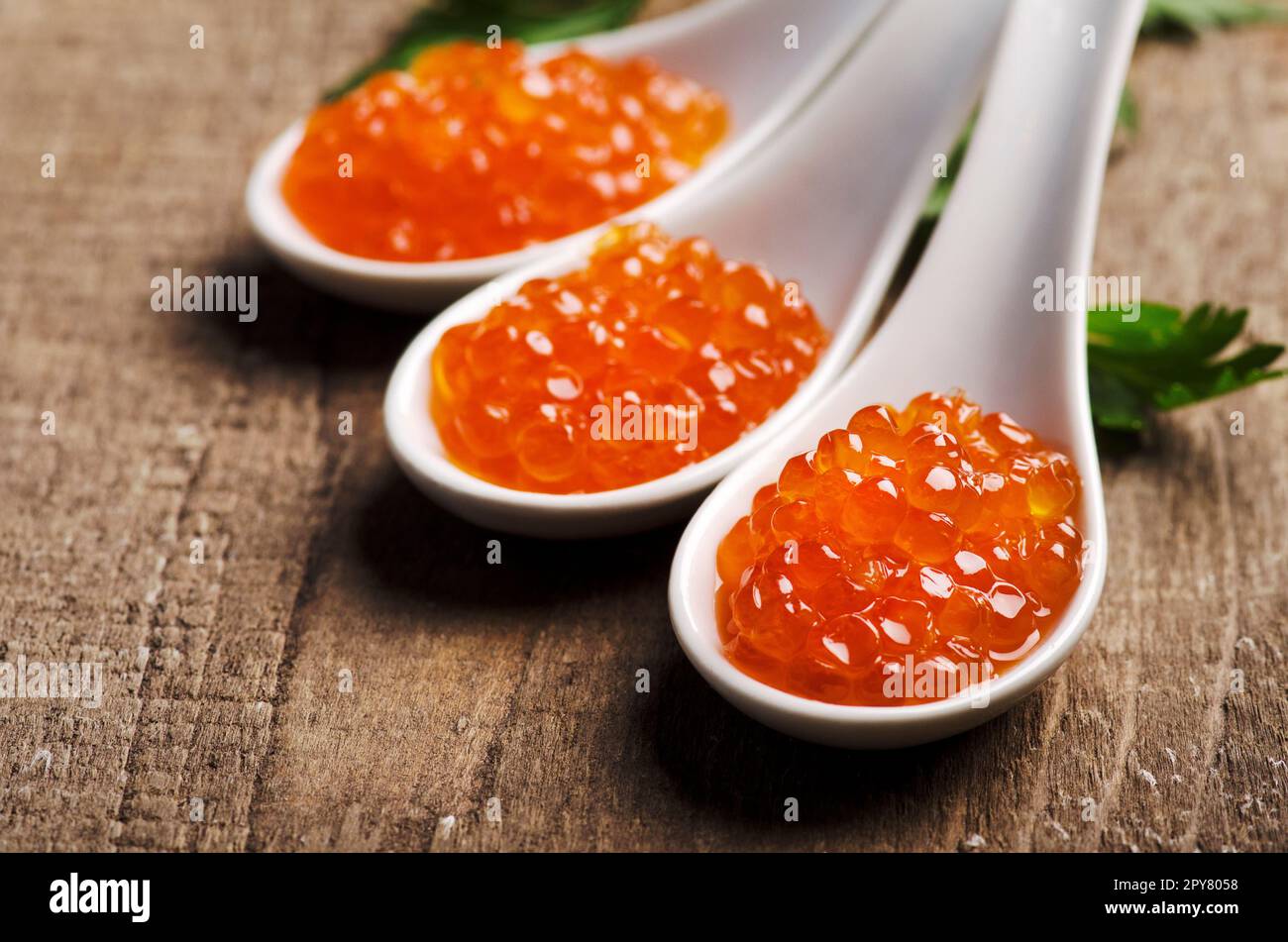 Primo piano del lussuoso caviale rosso in cucchiaio bianco con prezzemolo su fondo di legno. Foto Stock