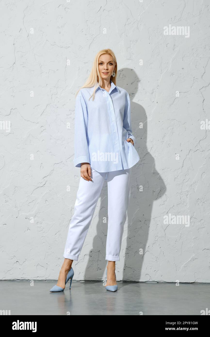 Donna con pantaloni bianchi, tacco a spillo e camicetta in cotone blu in posa vicino a una parete bianca Foto Stock