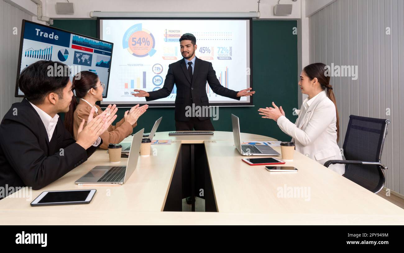 Uomo d'affari in abito nero che presenta i suoi prodotti con un grande monitor digitale. Riunione del team dei dirigenti aziendali in un ufficio moderno con computer portatile, tablet, telefono cellulare e caffè sul tavolo. Foto Stock