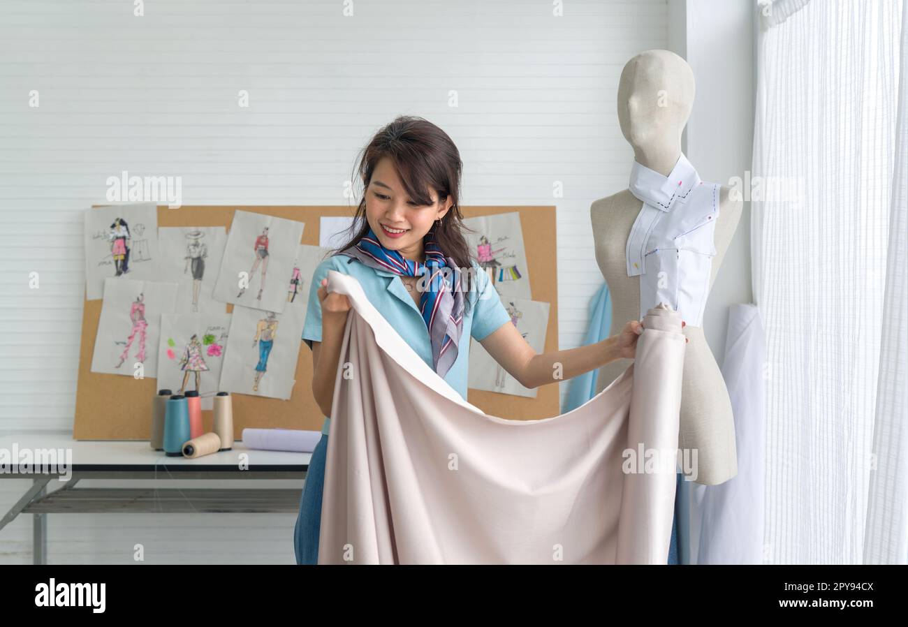 Giovane comò asiatico in sciarpa a righe verificare la qualità del tessuto beige fine dal rotolo. Camera di stilista. Foto Stock