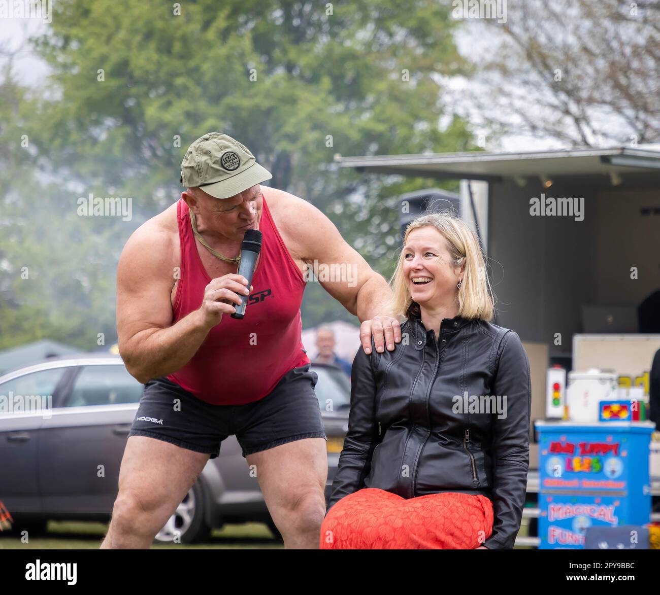 Culcheth, Regno Unito. Lunedì, 1 maggio, 2023, Culcheth, Inghilterra - nonostante i cieli grigi e alcune piovose, Culcheth e Glazebury Community Day sul verde del villaggio che offre intrattenimento da parte del Children’s Entertainer Ian “Zippy” Lees, Northern Stars Dance & Fitness e il Mighty Smith Strongman Show, con sidestalls e luna Park. Adrian Smith parla con una donna seduta su una sedia prima di sollevarla in aria credito: John Hopkins/Alamy Live News Foto Stock