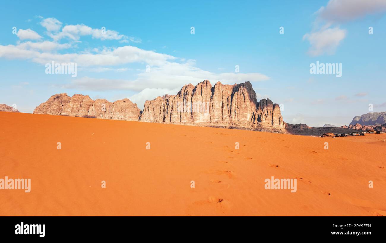 Deserto di sabbia arancione, formazioni rocciose e montagne sullo sfondo, cielo blu sopra, tende da campeggio visibili a distanza - scenario tipico a Wadi Rum, Giordania Foto Stock
