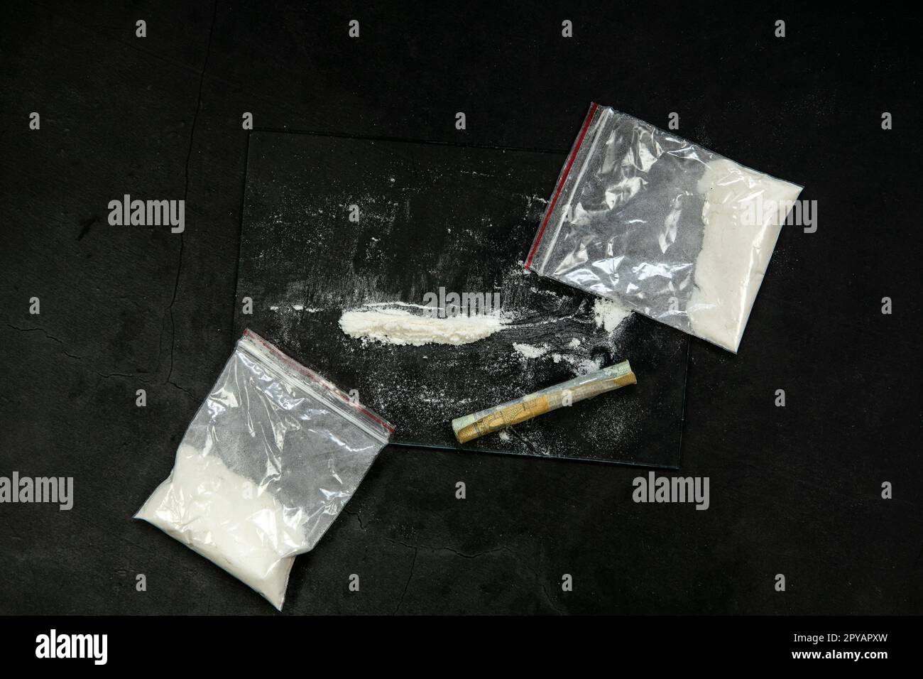 Linee di polvere di cocaina, banconote arrotolate e farmaci in tasca in busta di plastica su fondo in vetro nero, vista dall'alto. Concetto di tossicodipendenza Foto Stock