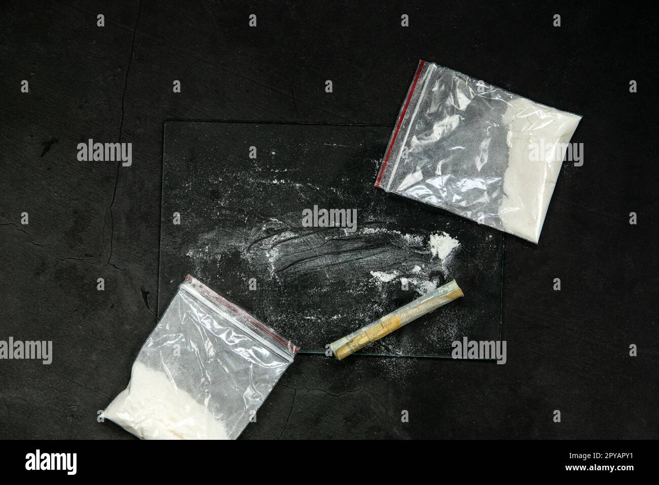 Linee di polvere di cocaina, banconote arrotolate e farmaci in tasca in busta di plastica su fondo in vetro nero, vista dall'alto. Concetto di tossicodipendenza Foto Stock