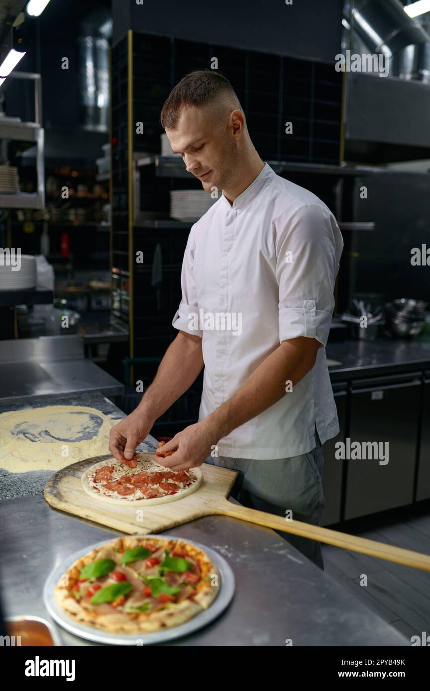 Pizzaiolo aggiungere prosciutto su base di pizza con formaggio, preparare piatto italiano Foto Stock