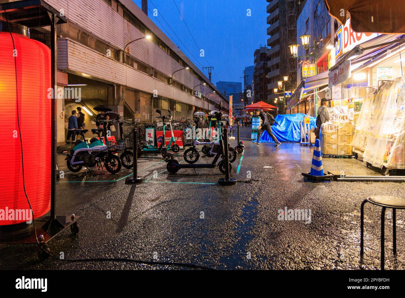 Osaka, Giappone - 29 aprile 2023: Scooter elettrici e biciclette parcheggiate da un luminoso negozio di droga e dalla stazione ferroviaria nella notte delle piogge Foto Stock