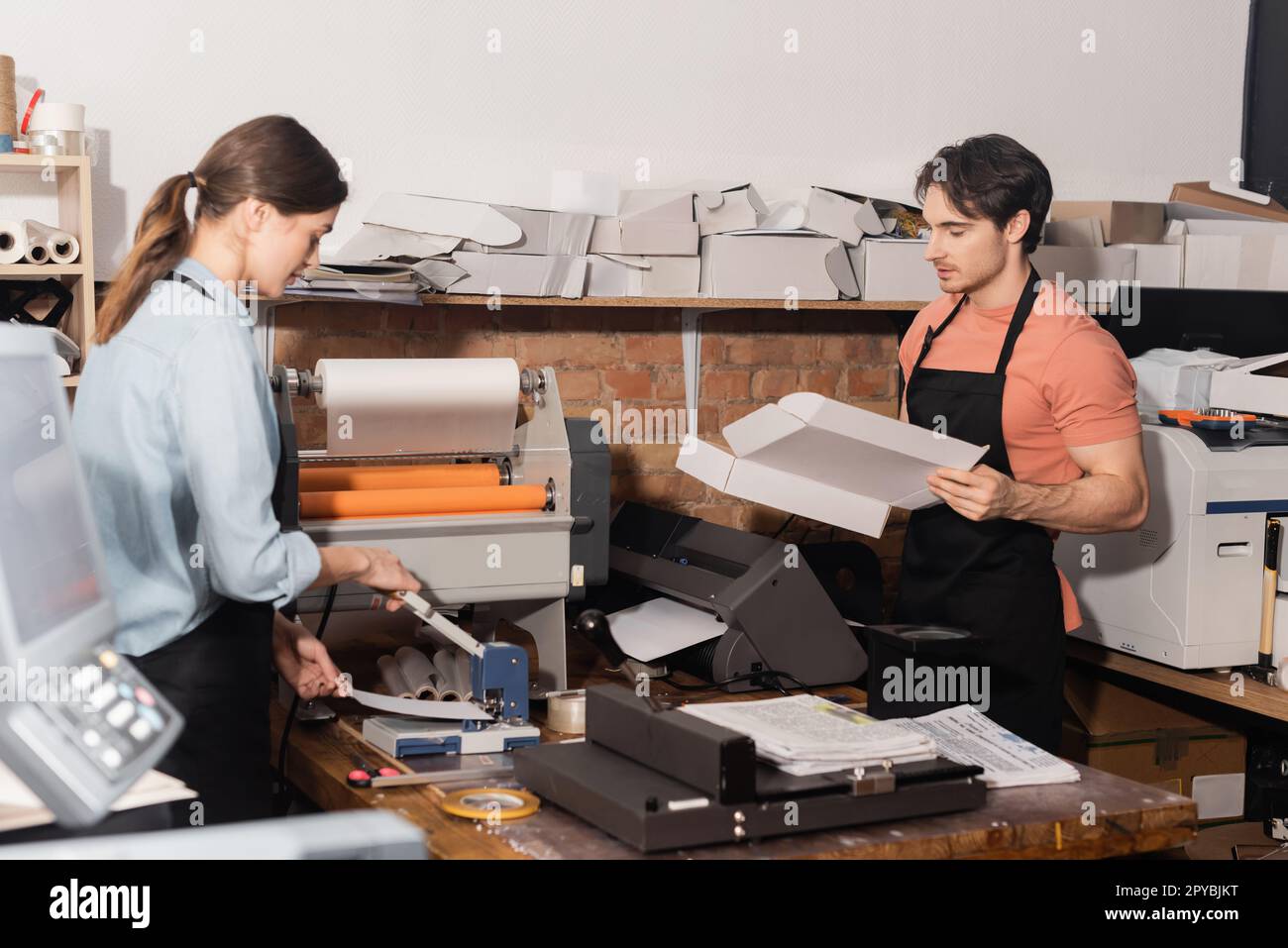 donna in grembiule che lavora con punzonatrice per carta accanto a collega con scatola di cartone, immagine stock Foto Stock