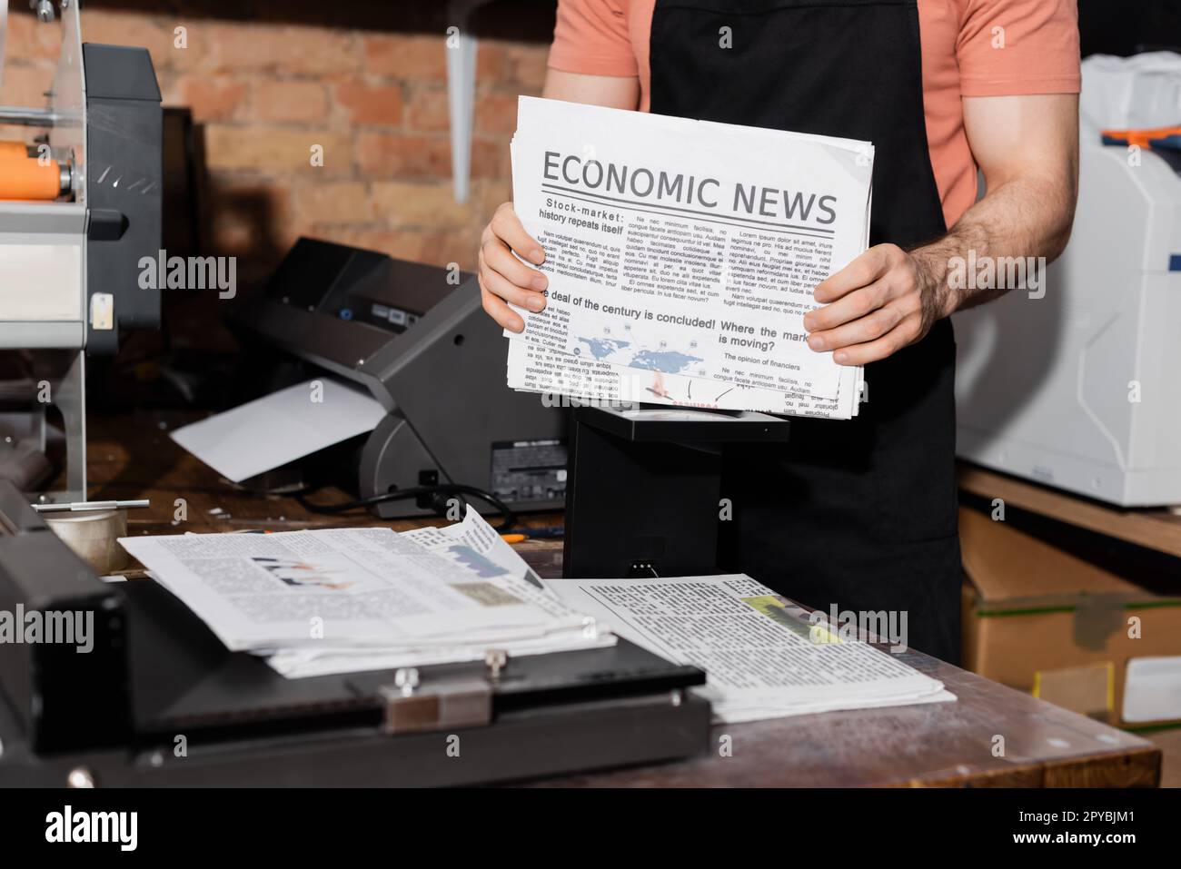 vista ritagliata del giovane tipografo in grembiule che tiene giornali con notizie economiche, immagine di riserva Foto Stock