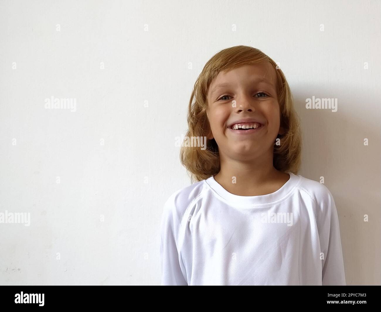 Una bambina di 6-7 anni con capelli chiari esprime gioia emotiva e un sorriso su sfondo bianco. Un bambino con pelle abbronzata. T-shirt bianca. Copia spazio Foto Stock