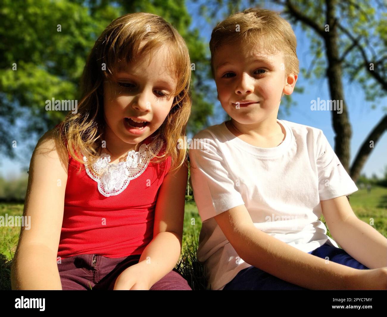 Due bambini sono seduti in un prato e parlano. Una ragazza di 6 anni e un ragazzo di 7 anni giocano all'aperto in un parco cittadino. Un ragazzo con una maglietta bianca, una ragazza con una maglietta rossa Foto Stock