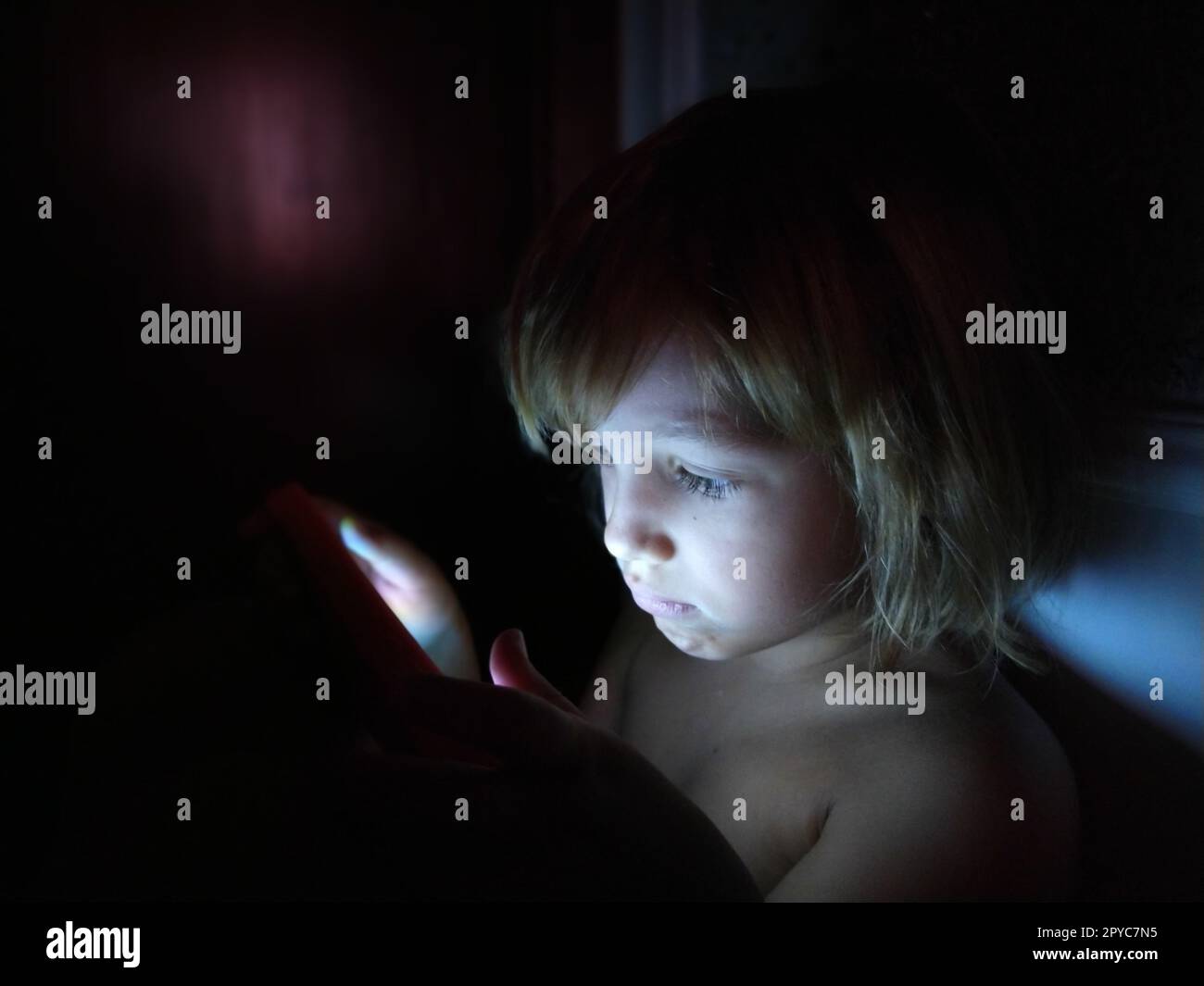 Una bambina carina gioca con un tablet. Un bambino di 7 anni con una faccia seria e capelli biondi guarda in un gadget. Copia spazio. Ragazza nella stanza. Parete scura e chiara. A casa di notte Foto Stock