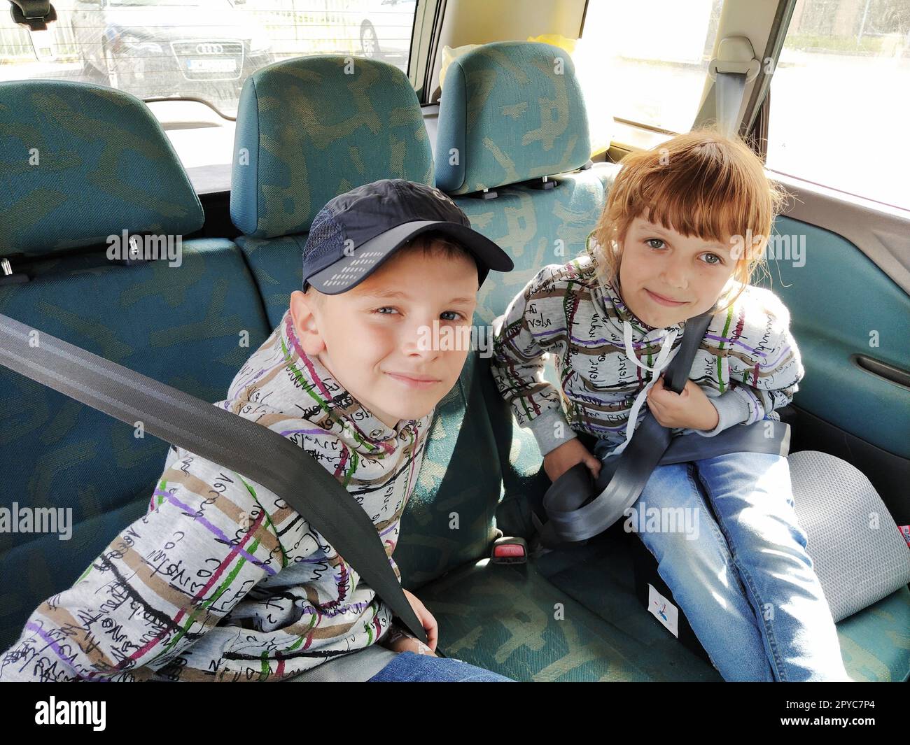 Bambini in macchina. Un ragazzo e una ragazza sono seduti sul sedile posteriore, ridono e lamentano. I bambini allacciano le cinture di sicurezza. Un bambino in berretto. Gemelli di facce diverse vestiti con gli stessi vestiti Foto Stock