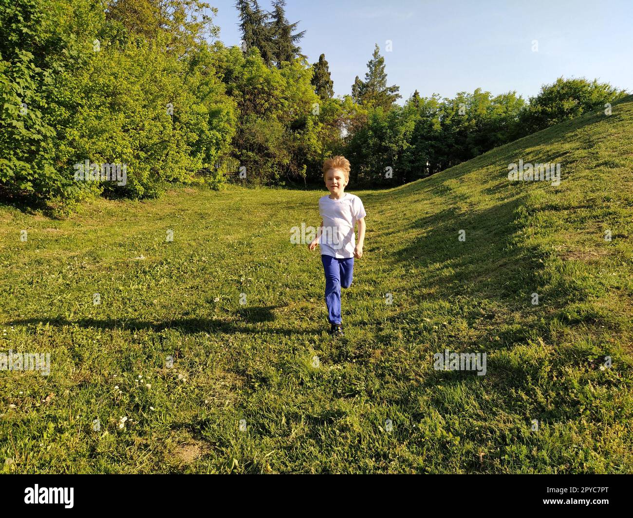 Un bambino corre nel parco. Un ragazzo con una T-shirt bianca e pantaloni blu. Prato o campo verde. Prato falciato. Cammina e gioca nella foresta. Attività sportive attive. Foto Stock